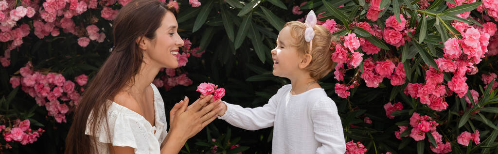 Vista lateral de la alegre niña dando flores a la madre cerca de la planta en Valencia, pancarta  - Foto, Imagen