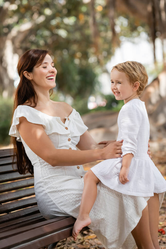 Cheerful toddler child in white dress sitting on mother in summer park  - Photo, image