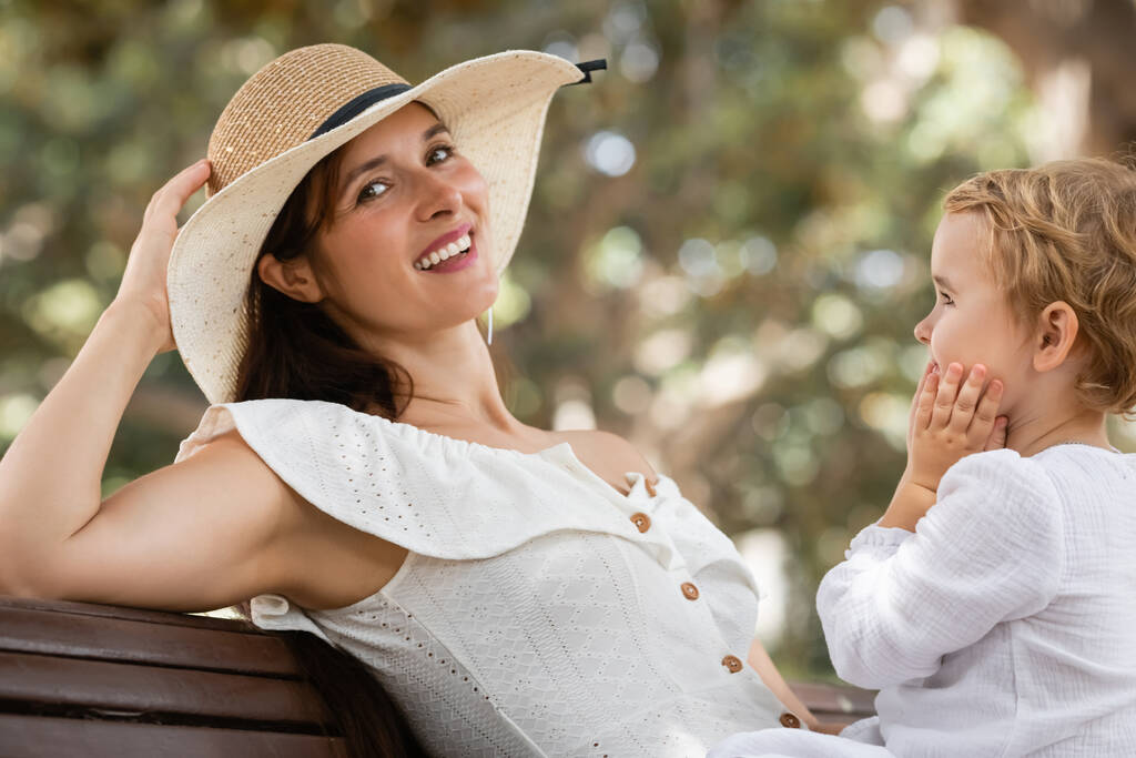 Усміхнена жінка в сонячному капелюсі і одягається, дивлячись на камеру біля дитини на лавці в парку
  - Фото, зображення