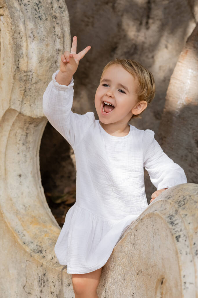 Excited toddler girl in dress showing peace sign on stone bench in Valencia  - Foto, Imagen