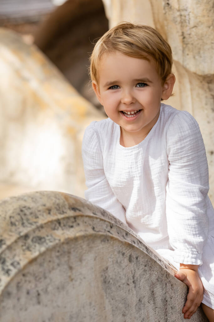 Усміхнена дівчинка в білій сукні сидить на кам'яній лавці в парку
  - Фото, зображення