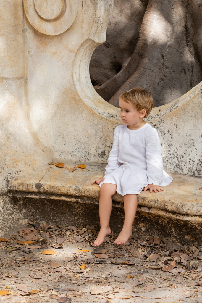 Barefoot baby girl in dress sitting on stone bench in park in Valencia  - Foto, immagini