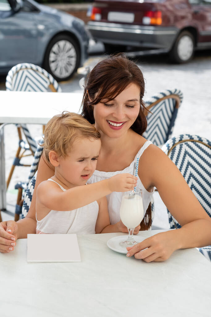 Lächelnde Frau betrachtet kleine Tochter mit Getränk in Outdoor-Café in Spanien  - Foto, Bild