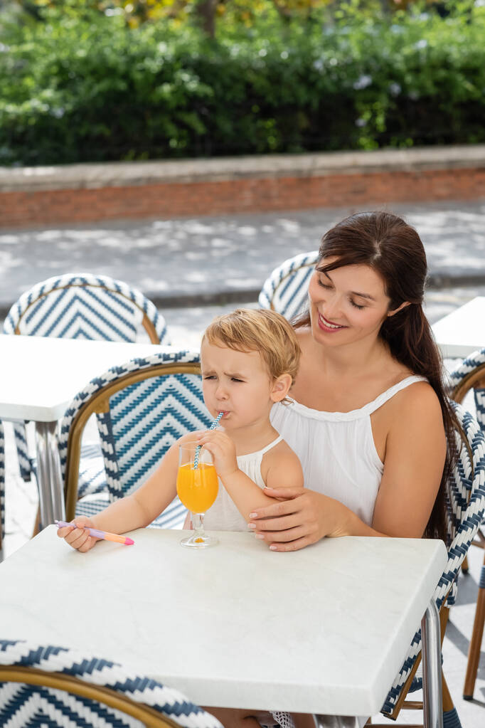 Cheerful mom looking at toddler girl drinking orange juice in outdoor cafe in Valencia  - 写真・画像