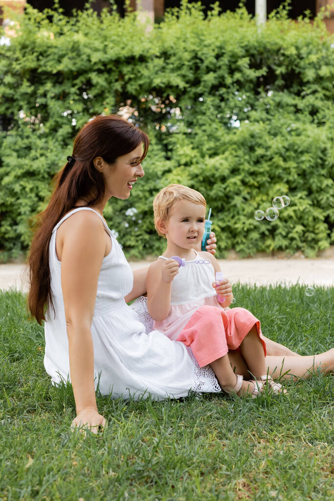 Usmívající se žena dívá na dceru s mýdlovými bublinkami na trávníku v letním parku  - Fotografie, Obrázek