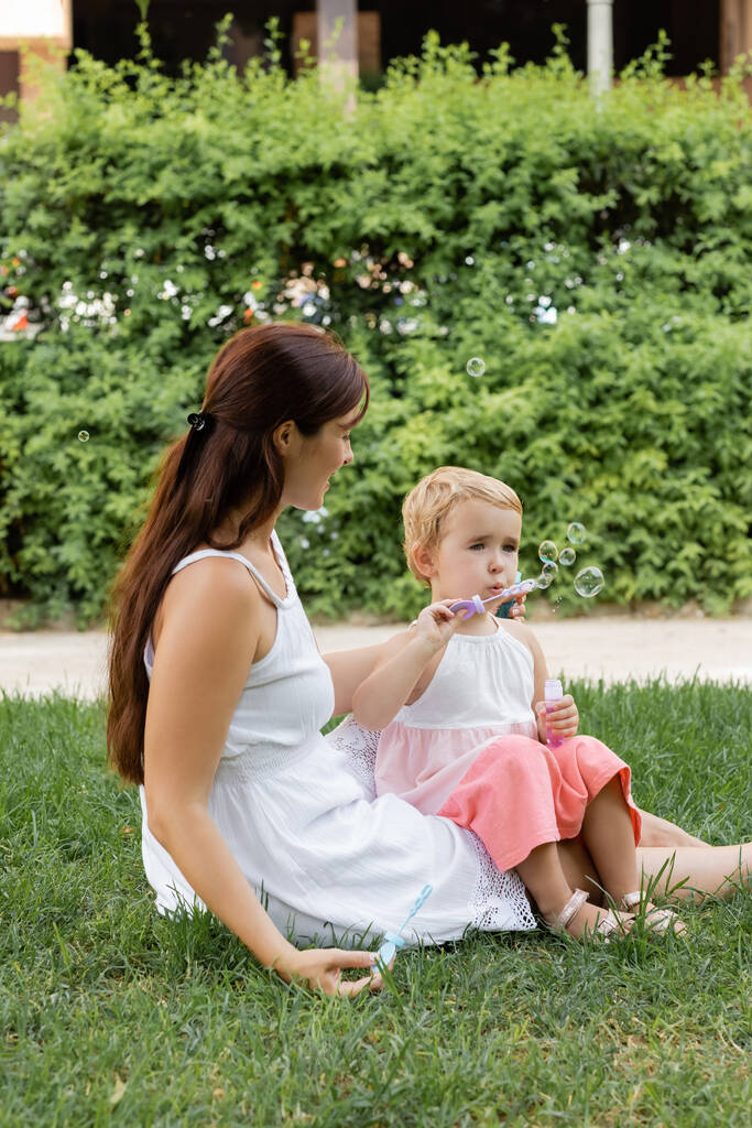 Nő nézi kisbaba lánya fúj szappanbuborékok a nyári parkban  - Fotó, kép