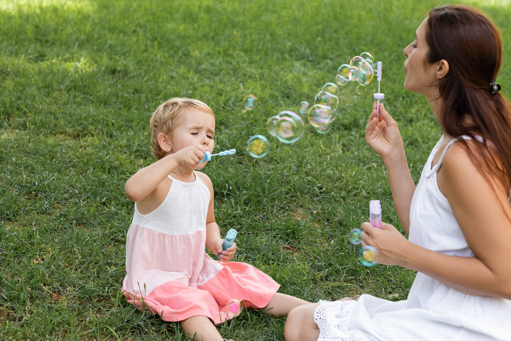 Daughter in dress blowing soap bubbles near mom on lawn in park  - 写真・画像
