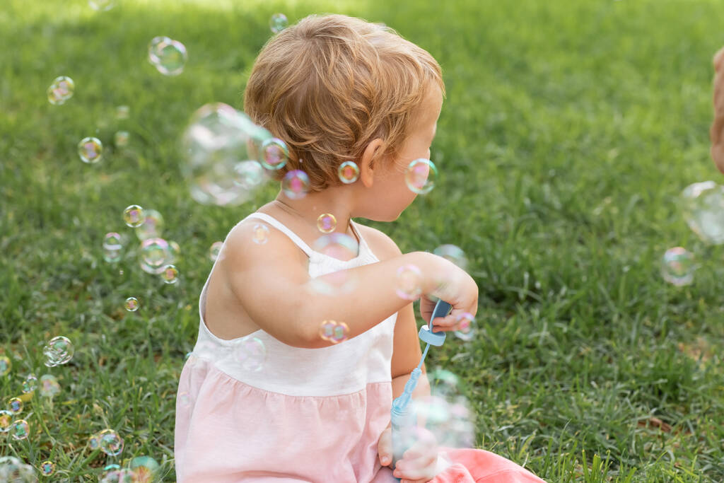 Zijaanzicht van baby meisje in jurk houden zeepbellen in de zomer park  - Foto, afbeelding