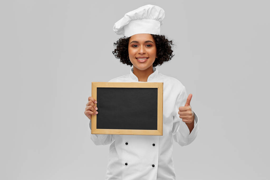cooking, advertisement and people concept - happy smiling female chef in toque holding chalkboard showing thumbs up over grey background - Photo, Image