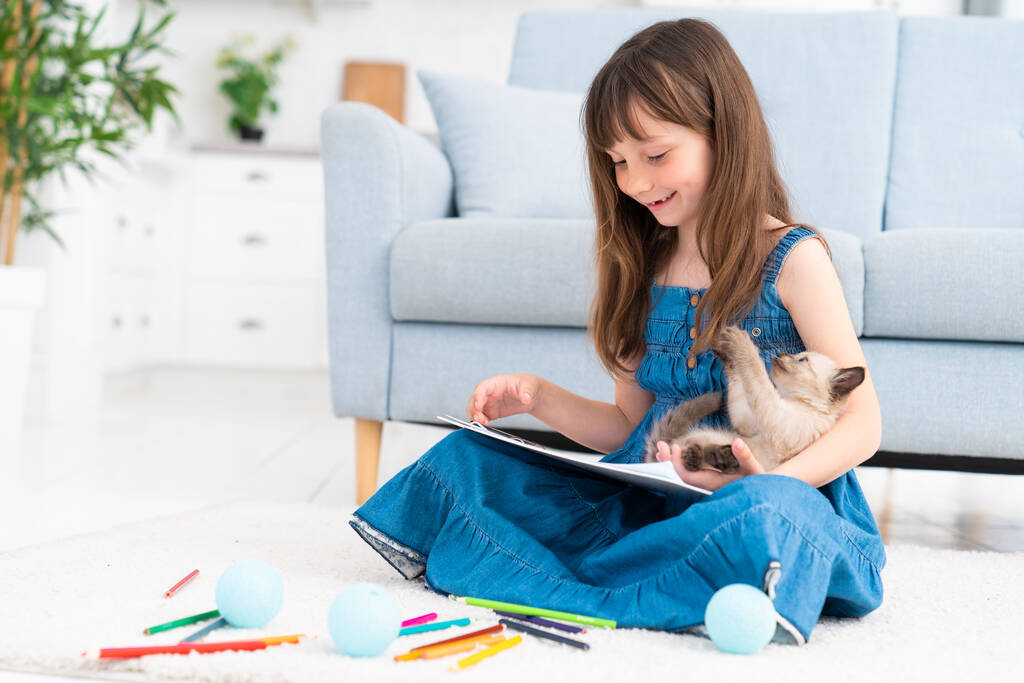 Egy aranyos lány otthon ül a padlón, színes ceruzákkal rajzol, és egy kiscicával játszik. A gyermekek fogalma és az állatok iránti szeretet. Kiváló minőségű fénykép - Fotó, kép