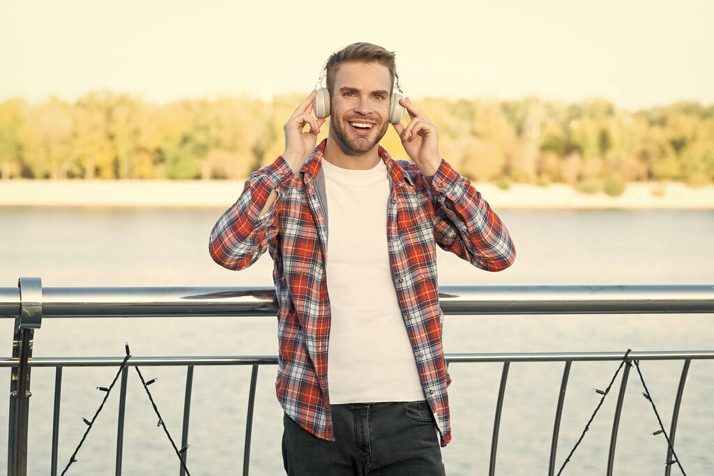 szczęśliwy młody modny mężczyzna moda model w słuchawki muzyczne i casual koszuli na zewnątrz, muzyka. - Zdjęcie, obraz