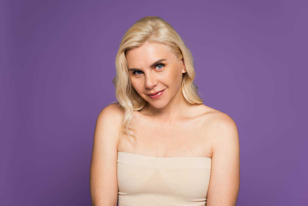 smiling blonde woman with bare shoulders looking at camera isolated on purple - Foto, imagen