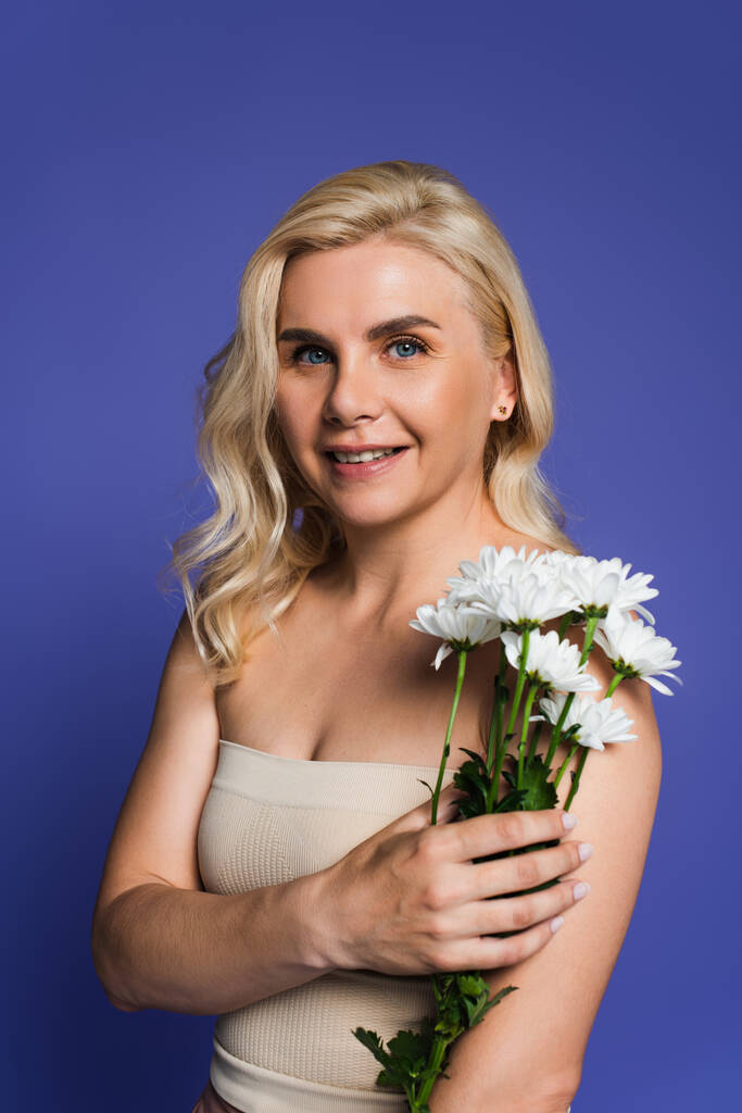 cheerful woman with bare shoulders looking at camera while holding flowers isolated on violet - Foto, Bild