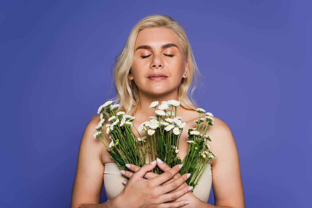 blonde woman with closed eyes smelling flowers isolated on purple - Photo, image