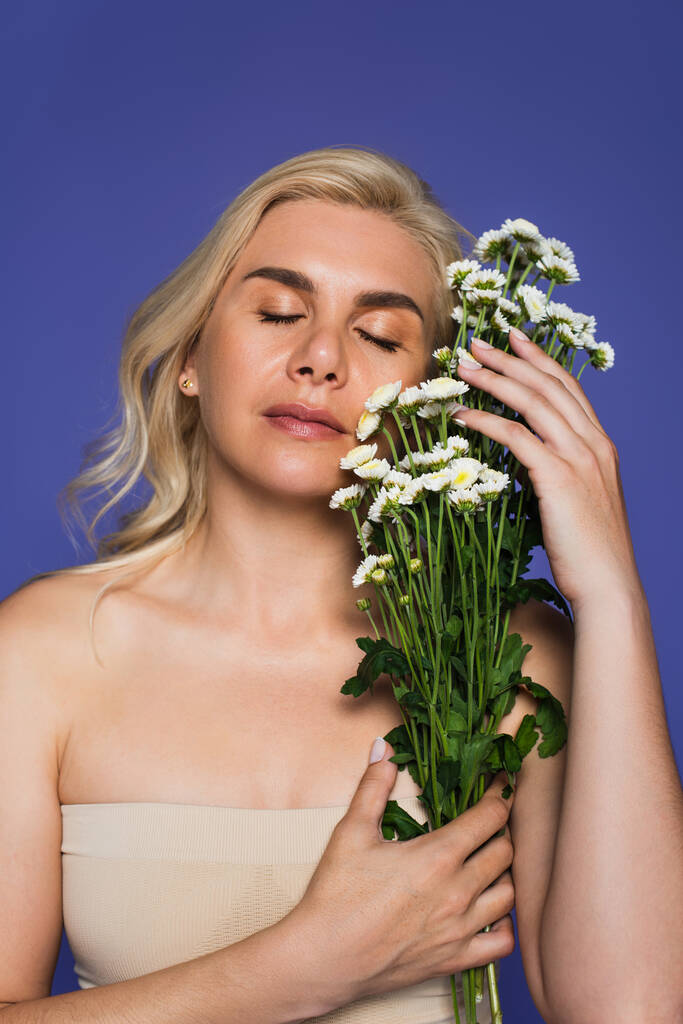 sinnliche und blonde Frau mit geschlossenen Augen, die Blumen isoliert auf Veilchen hält - Foto, Bild