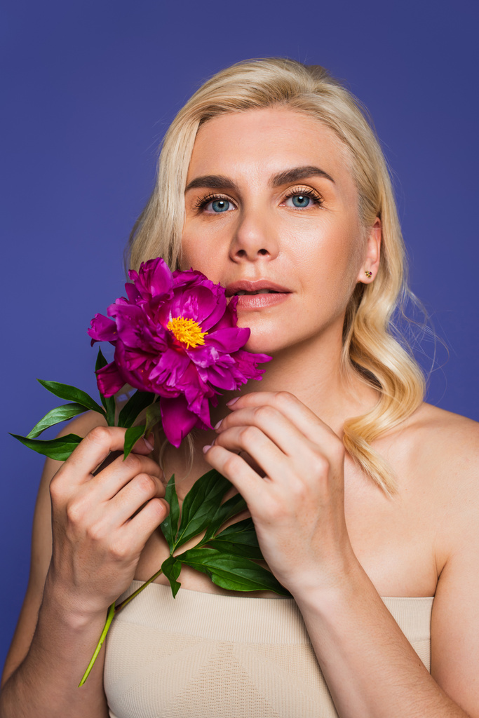 blonde woman with blue eyes holding blooming flower and looking at camera isolated on purple - Photo, Image