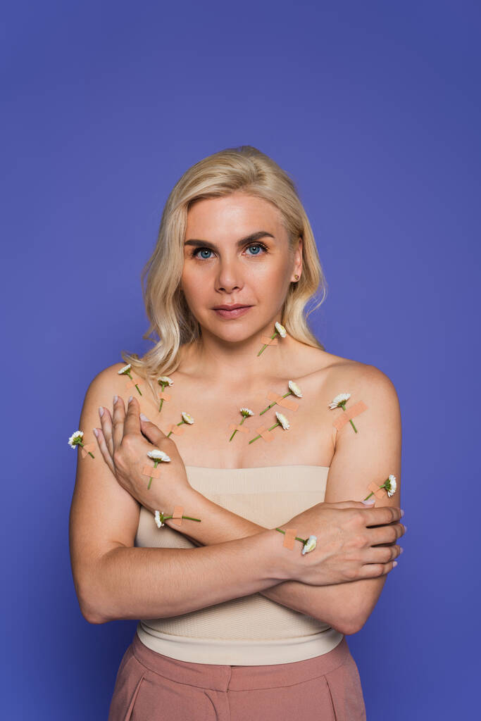 blonde woman with plasters and fresh flowers on body looking at camera isolated on purple - Φωτογραφία, εικόνα
