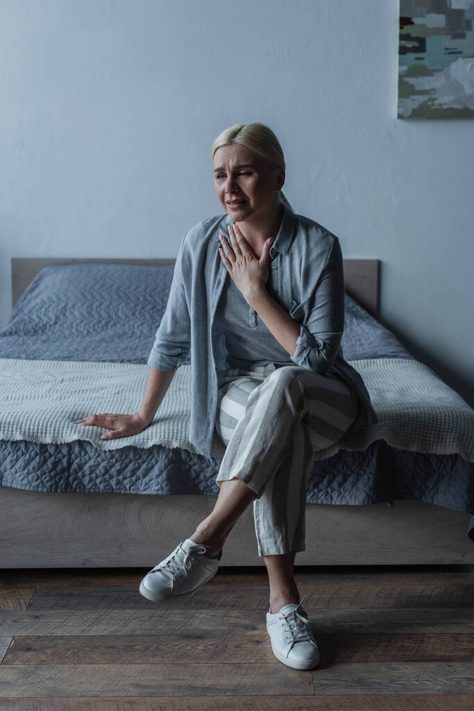 full length of upset blonde woman with menopause sitting on bed and touching chest - Foto, imagen