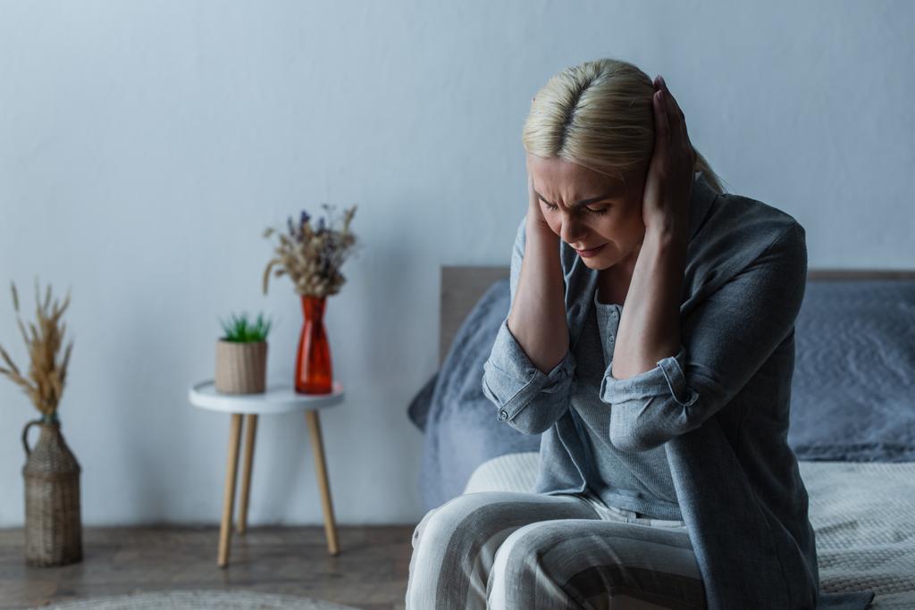 Menopozlu yorgun bir kadın yatak odasında otururken baş ağrısı çekiyor. - Fotoğraf, Görsel
