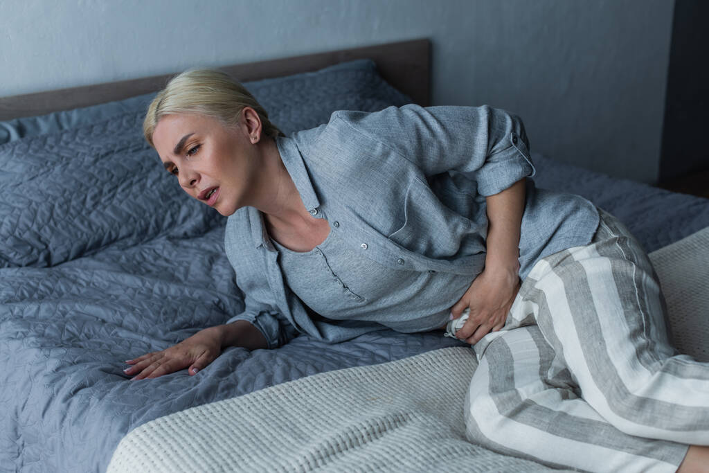 tired blonde woman with menopause suffering from abdominal pain in bedroom - Photo, Image