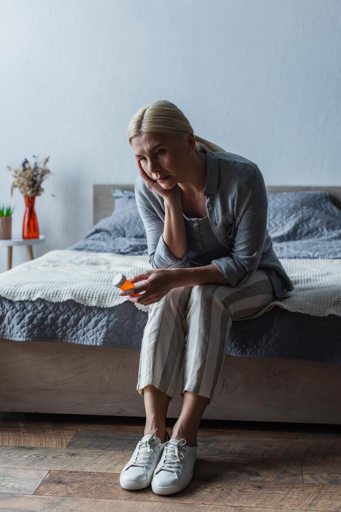 upset blonde woman with menopause holding bottle with pills and sitting on bed - Fotografie, Obrázek