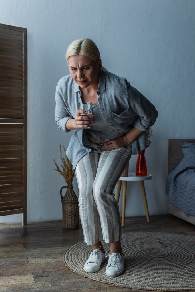stressed woman with menopause holding glass of water while suffering from abdominal pain - Foto, Imagen