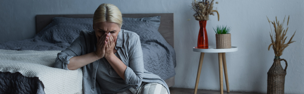 mulher com menopausa sentado perto da cama enquanto cobre o rosto e sofrendo de dor abdominal, banner - Foto, Imagem