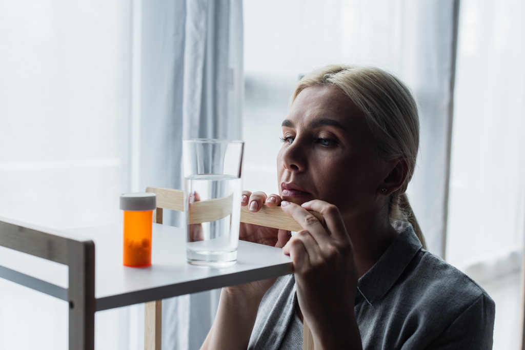 blonde woman with menopause looking at medication and glass of water on table - Fotó, kép