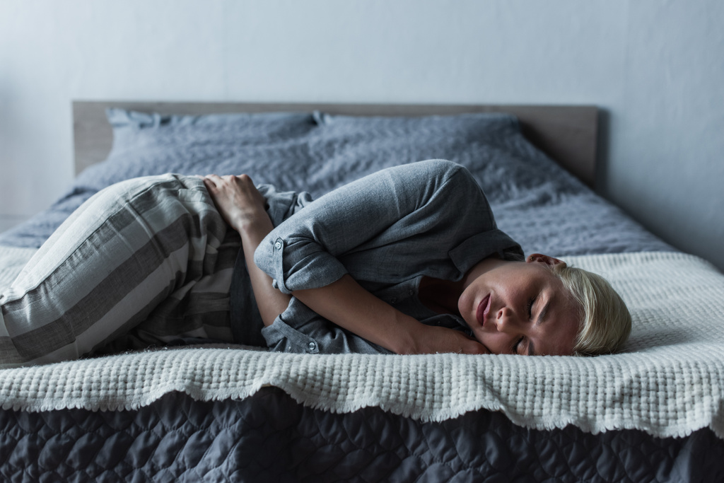 blonde woman lying on bed and suffering from stomach ache during menopause  - Foto, afbeelding