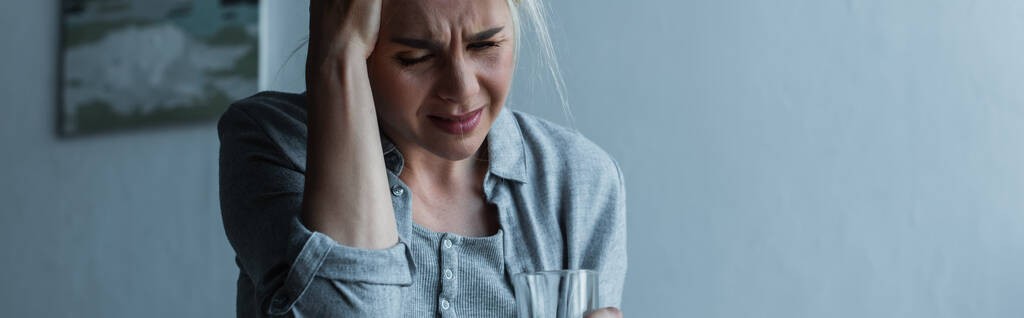 uitgeput blonde vrouw die glas vasthoudt terwijl ze migraine heeft tijdens de menopauze, spandoek - Foto, afbeelding