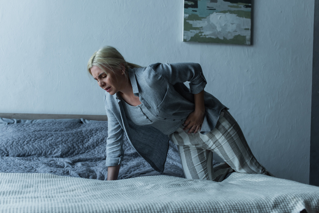 blonde woman getting into bed while feeling pain in stomach during menopause  - Fotó, kép