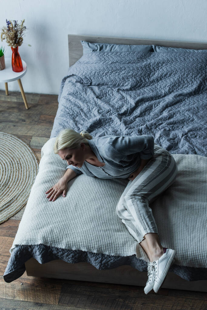 vista de ángulo alto de la mujer frustrada acostada en la cama mientras siente dolor en el estómago durante la menopausia  - Foto, Imagen