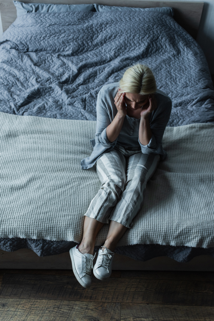 wysoki kąt widzenia blondynka siedzi na łóżku podczas bólu głowy podczas menopauzy  - Zdjęcie, obraz