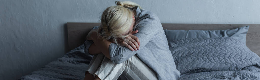 blonde woman covering face and hugging knees on bed while feeling pain in stomach during menopause, banner - Foto, afbeelding