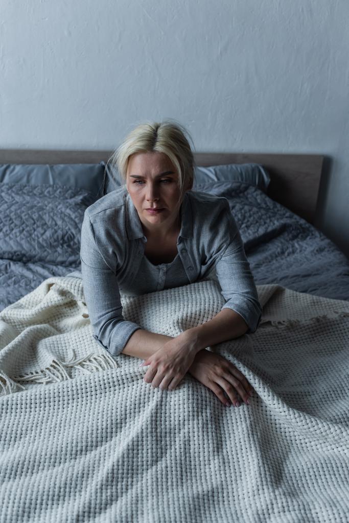 mulher loira e deprimida com olhos azuis sentindo-se mal durante a menopausa enquanto está sentada na cama - Foto, Imagem