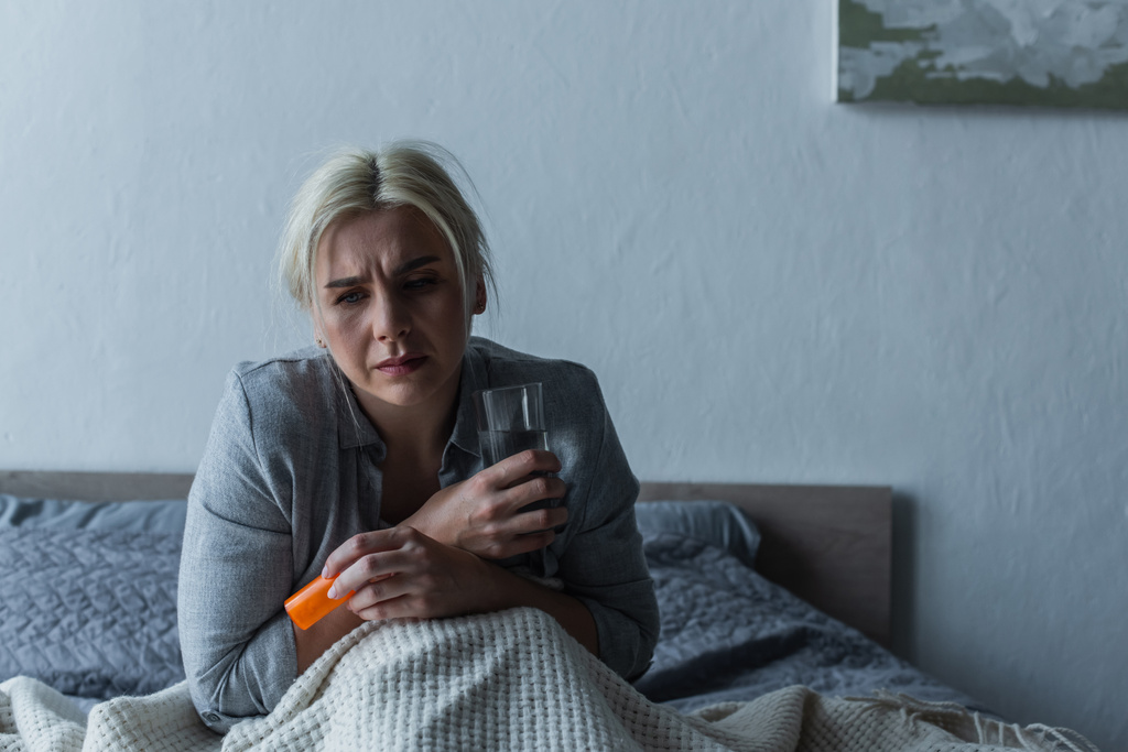 истощенная женщина с кульминацией сидя в постели, держа стакан воды и болеутоляющие  - Фото, изображение