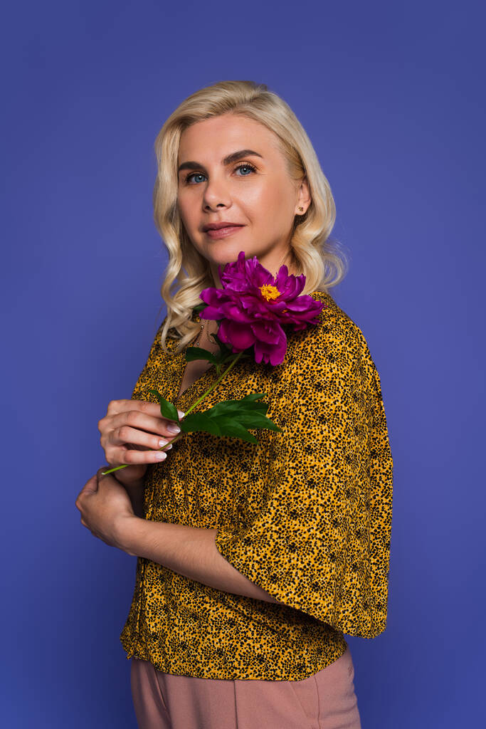 blonde woman with blue eyes holding flower with green leaves isolated on violet - Foto, afbeelding