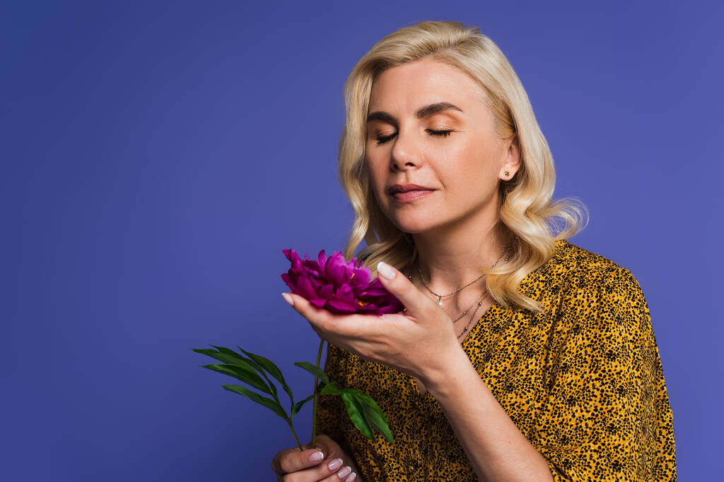 blonde woman in blouse smelling purple flower with green leaves isolated on violet - Φωτογραφία, εικόνα