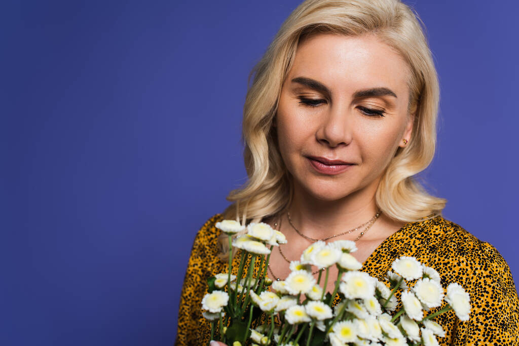 blonde woman in blouse looking at bouquet of white flowers isolated on violet - Fotoğraf, Görsel