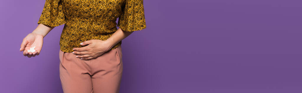 bijgesneden beeld van vrouw met buikpijn holding pillen geïsoleerd op paars, banner - Foto, afbeelding