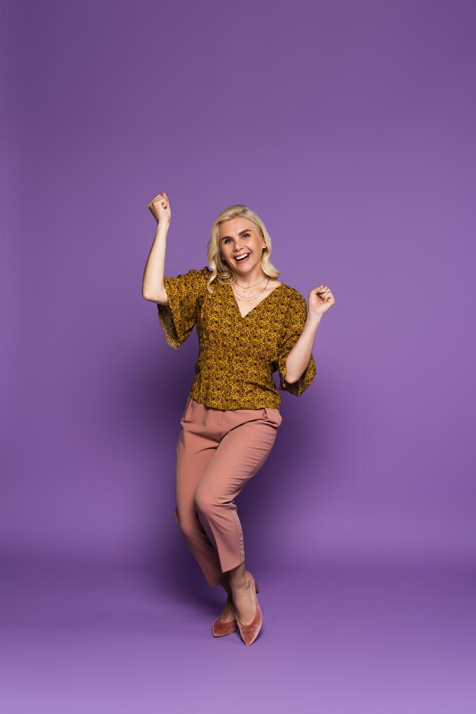 полная длина счастливой блондинки в блузке с менопаузой и ликованием на фиолетовом - Фото, изображение