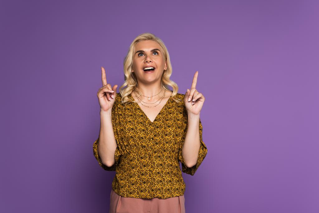 femme blonde étonnée avec ménopause souriante et pointant vers le haut avec les doigts isolés sur violet - Photo, image