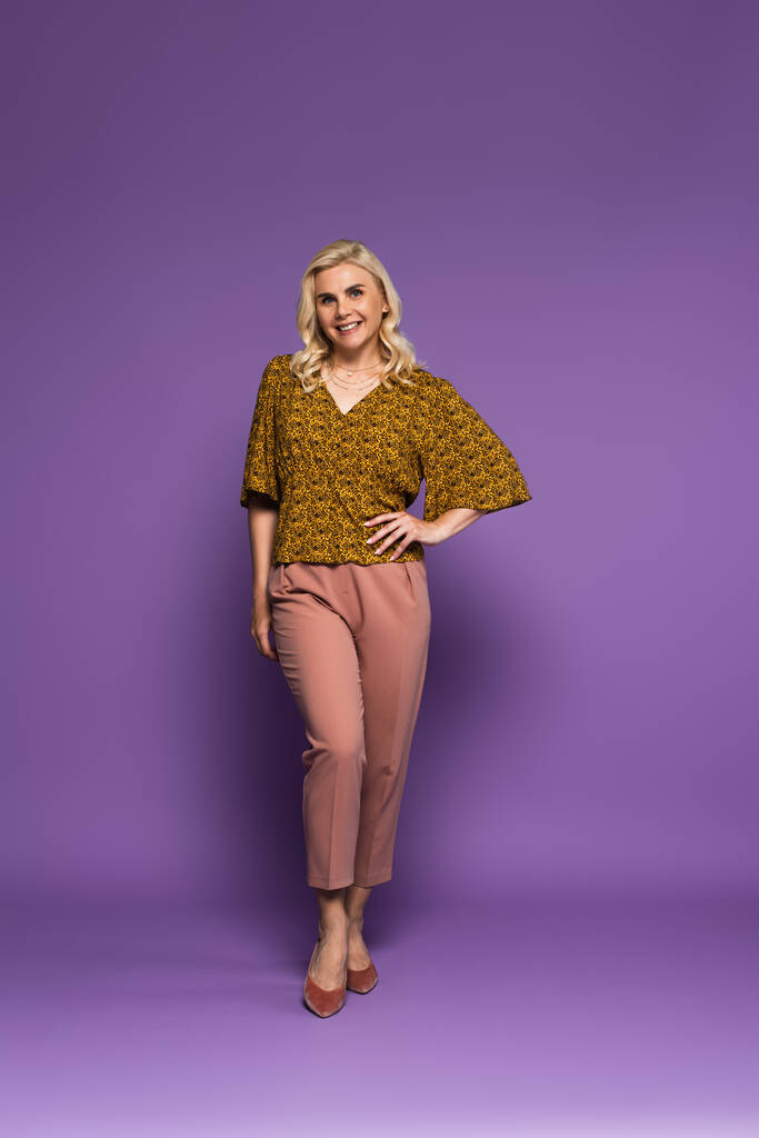 full length of blonde woman with menopause posing with hand on hip and smiling on purple  - Foto, Imagem