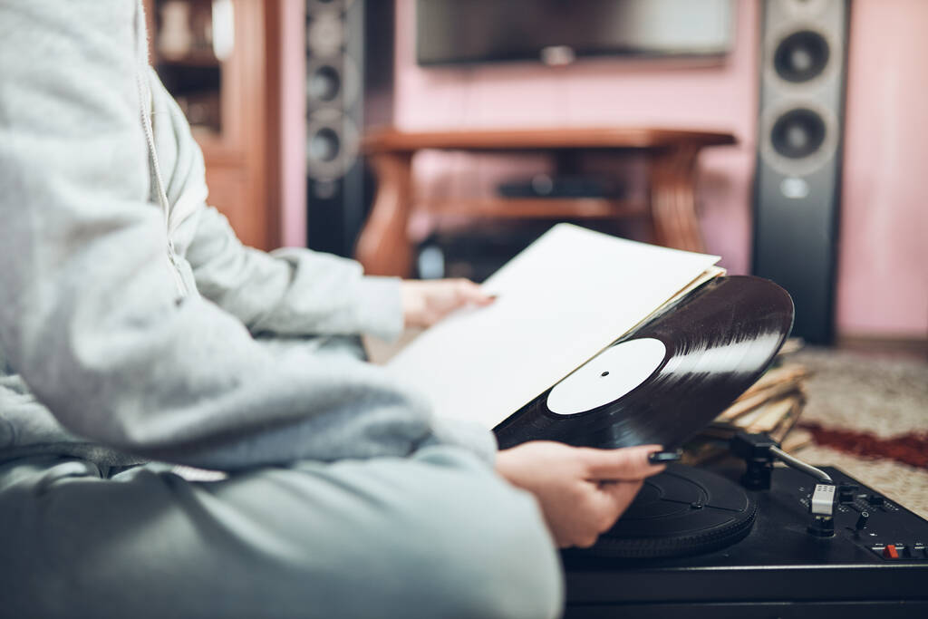 Νεαρή γυναίκα που ακούει μουσική από πικάπ βινυλίου. Ρετρό και vintage στυλ μουσικής. Κορίτσι κρατώντας αναλογικό δίσκο κάθεται στο δωμάτιο στο σπίτι. Γυναίκα απολαμβάνοντας μουσική από την παλιά συλλογή δίσκων - Φωτογραφία, εικόνα