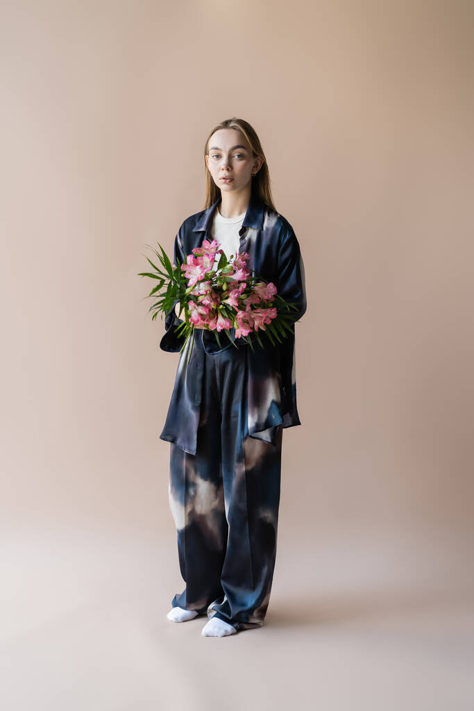 full length of woman in tie-dye style clothes standing with floral bouquet on beige background - Valokuva, kuva
