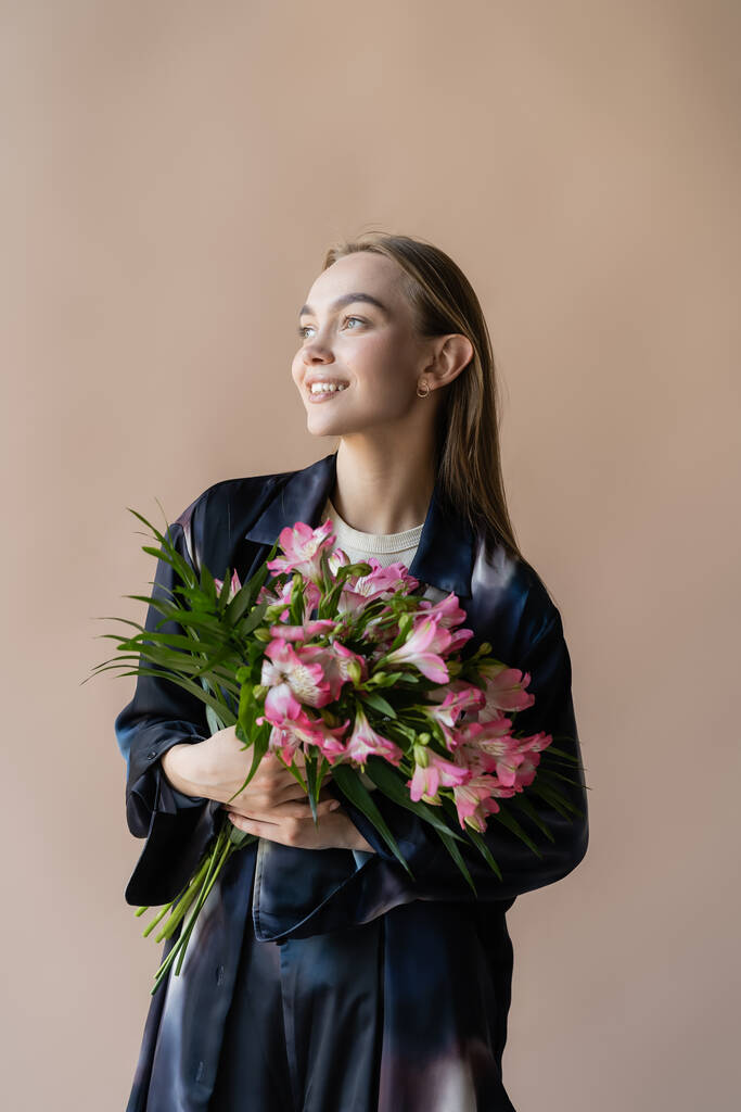 donna felice e alla moda con bouquet floreale fresco guardando altrove isolato sul beige - Foto, immagini