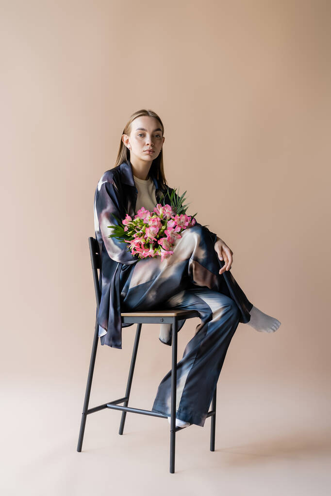 full length of stylish woman with alstroemeria bouquet posing on chair on beige background - Valokuva, kuva