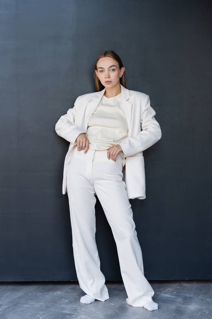full length of woman in white suit and socks standing with hands on waist on black background - Foto, afbeelding