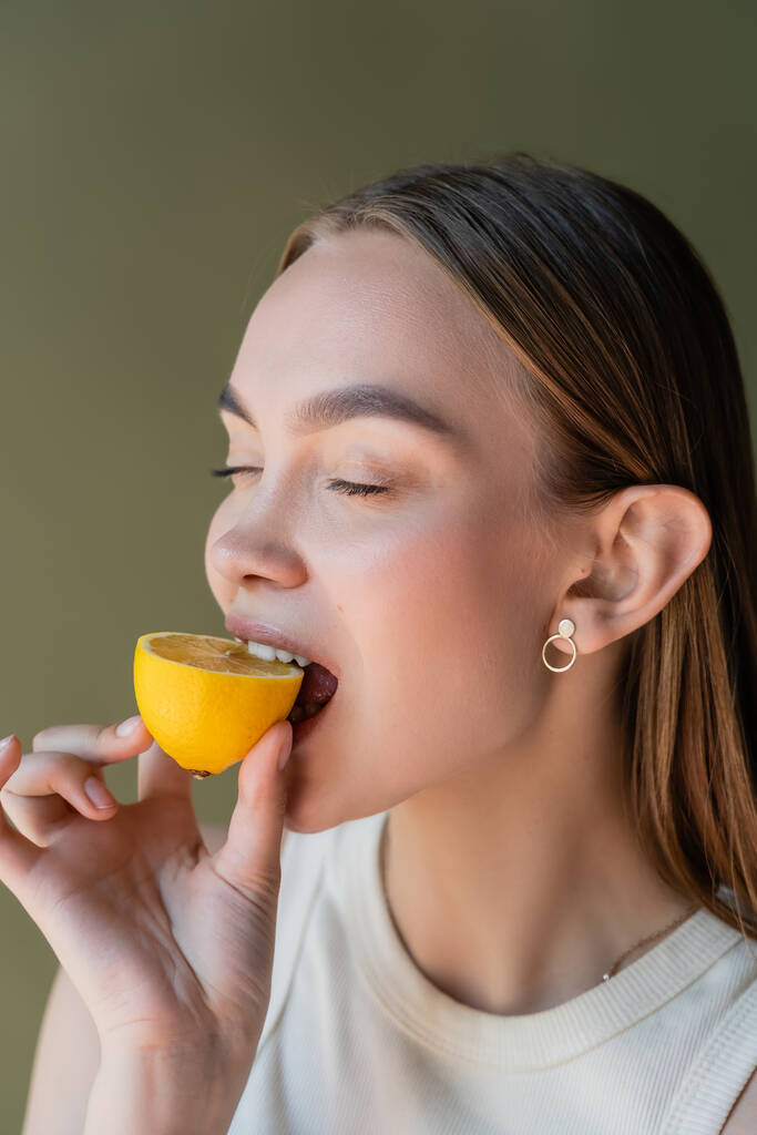 young woman with closed eyes biting juicy lemon isolated on green - Foto, imagen