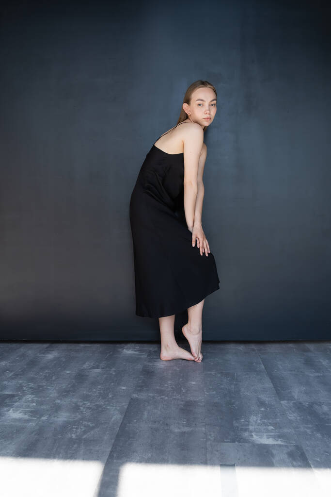full length of slim barefoot woman in black strap dress near dark wall - Foto, Imagen